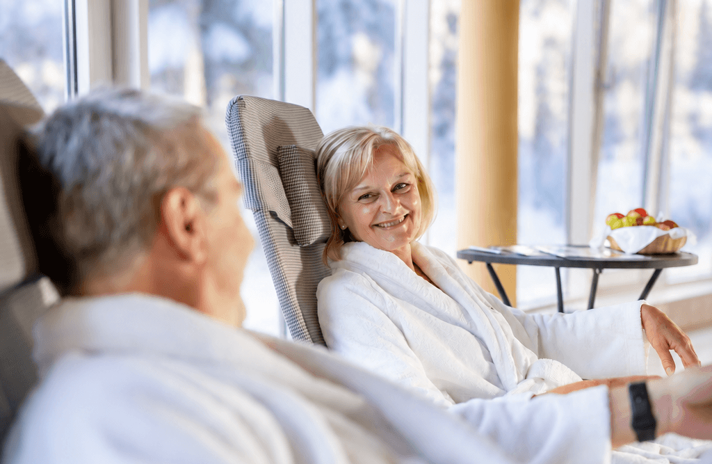 Entspannung im winterlichen Spa-Lounge Bereich_quer Zwei Personen in weißen Bademänteln entspannen auf bequemen Liegen in einem lichtdurchfluteten Spa-Ruhebereich. Eine lächelnde Frau blickt zu ihrem Begleiter. Durch die großen Panoramafenster ist eine verschneite Winterlandschaft zu sehen.