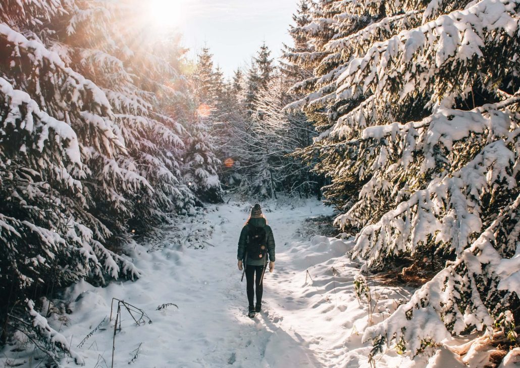 Winterwanderung_9609 Eine Person mit Winterjacke, Haube und Rucksack wandert auf einem verschneiten Waldweg durch einen winterlichen Nadelwald. Sonnenstrahlen fallen durch die schneebedeckten Bäume und tauchen die Winterlandschaft in warmes Licht.